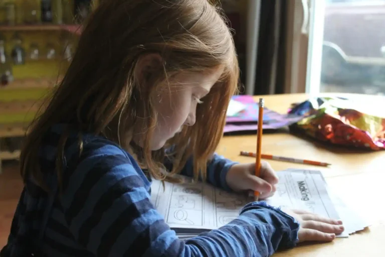 Young girl seated at a desk by a window, carefully writing answers on a worksheet with a pencil