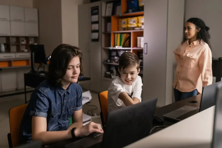Students working together on laptop computers in a classroom setting, guided by a teacher, exploring technology and learning